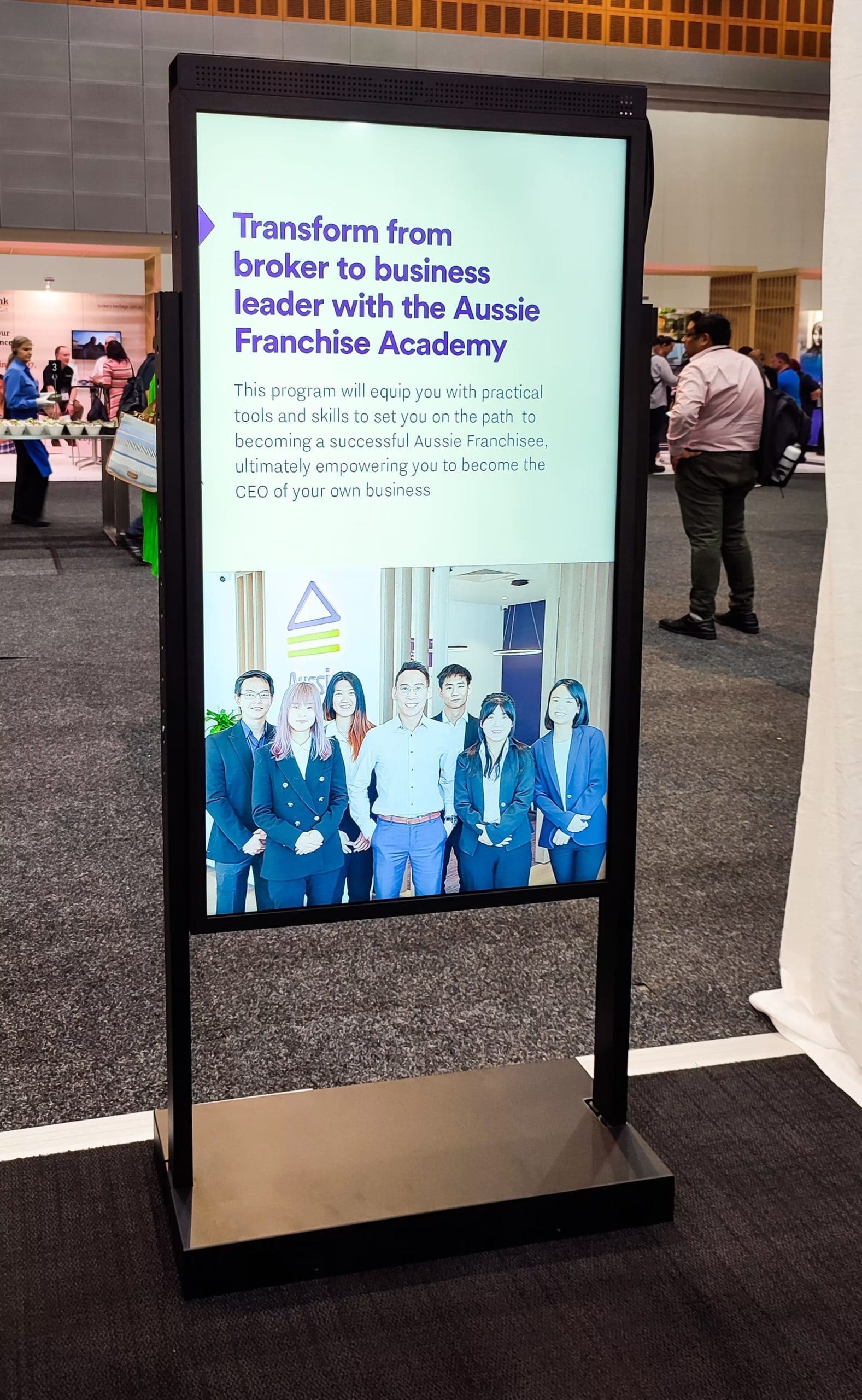 Vertical digital kiosk at a business expo promoting the Aussie Franchise Academy with group photo of team members.