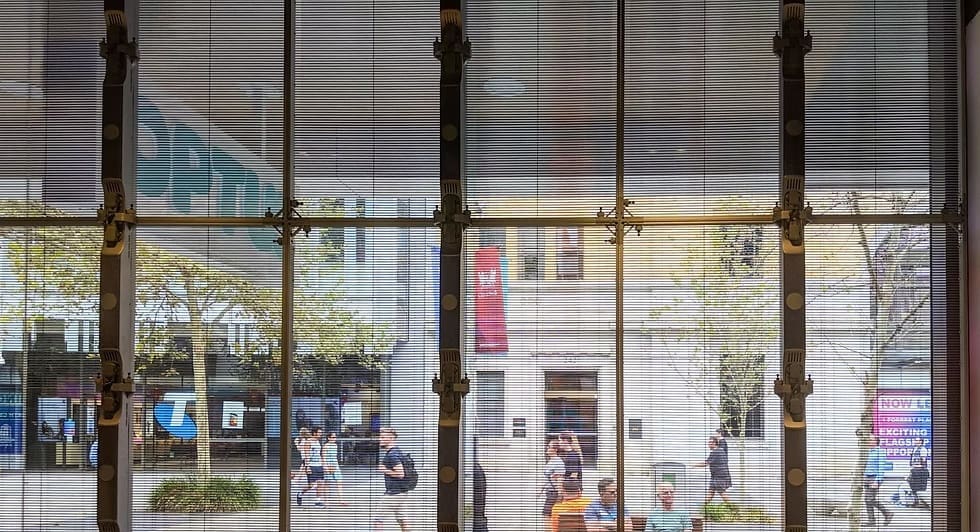 Transparent LED display wall viewed from inside the Optus store, showing street pedestrians and nearby shops.